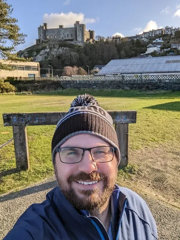 Jamie Clarke, smiling in a woolly hat on a sunny day out, with a castle on the hill behind him.
